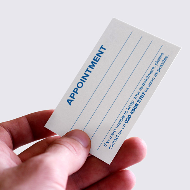 Custom printed appointment card by TradePrintingUK.com, held in a hand against a light gray background, showing the front with bold APPOINTMENT heading in blue, blank lines for client name, date, and details, plus cancellation instructions to contact the London telephone number as soon as possible, ideal for salons, clinics, or service businesses.
