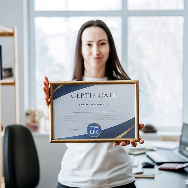 Woman in a white top holding a framed custom printed award certificate by TradePrintingUK.com, featuring TPUK logo, blue wavy border, and Certificate Proudly Presented To text, displayed in a bright modern office with a window, desk, and bookshelves in the background.