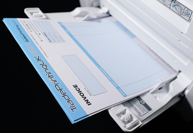 Custom printed invoice on 100gsm Bond by TradePrintingUK.com loaded in a laser printer tray. The personalised business form features a light blue header with geometric design and invoice fields, demonstrating jam-free paper performance for high-volume office printing