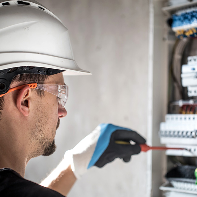 UK electrician in white hard hat and safety glasses inspects consumer unit circuit breakers and wiring in domestic property, demonstrating electrical testing and safety compliance work requiring professional receipts and certification documentation.
