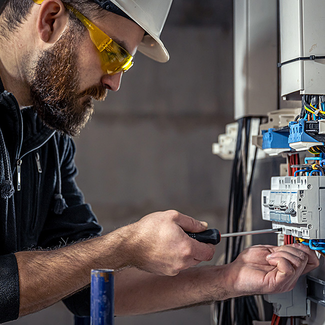 Electrician wearing hard hat and yellow safety glasses tightens consumer unit terminals with screwdriver, working on circuit breakers and wiring for UK electrical installation and safety compliance.