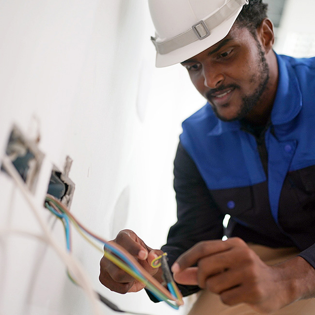 UK electrician in white hard hat and blue workwear strips and prepares electrical cable wiring for socket installation in domestic property, demonstrating professional electrical work requiring job receipts and documentation.