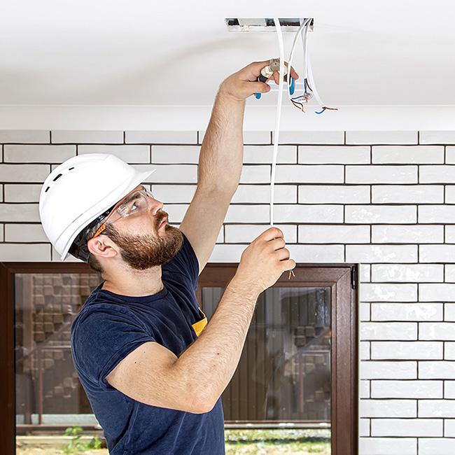 UK electrician in hard hat wiring ceiling fixture, demonstrating electrical isolation and safety compliance for trade contractors.