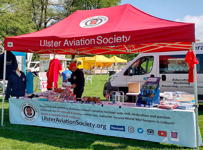 Long custom printed PVC banner by TradePrintingUK.com on the Ulster Aviation Society red pop-up tent at the Lord Mayors Show event in Lisburn, promoting 'Checkout our Northern Irelands heritage with aircraft and Second World War hangars in Lisburn', with Facebook, Twitter, Instagram icons, alongside a shop table with books and flyers on grassy grounds with yellow tents and their crew white van nearby.