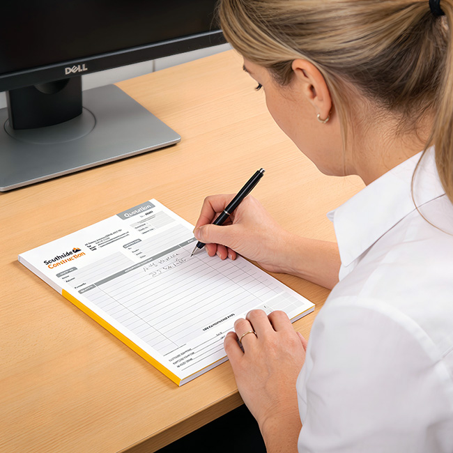 Professional office manager Woman with blonde hair writing on a custom printed carbonless quotation pad from TradePrintingUK.com at an office desk. The personalised NCR form features custom sequential numbering, and a company logo with bespoke address, ideal creating quotations for customers for goods and services. A computer monitor is in the background.