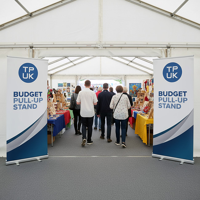 Two TradePrintingUK.com budget roller banner stands with blue TPUK branding, prominently placed at the entrance of a busy white marquee at a local market, demonstrating effective, high-visibility event signage for small businesses and stalls.