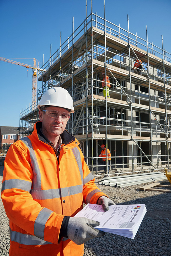Construction worker in a white hard hat and orange hi-vis jacket holding a stack of scaffolding handover certificate forms on a building site, with scaffolded structure and other workers in the background.