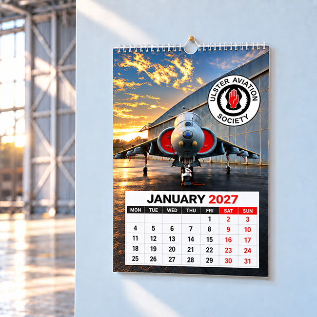 A3 charity fundraising wall calendar displayed inside an aviation heritage society hangar, with aircraft softly visible in the background.